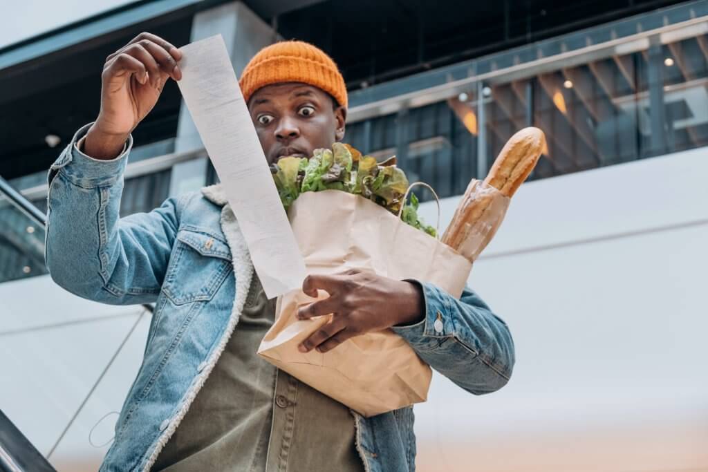 Doubting African-American person in denim jacket looks at sales paper receipt total holding pack with food products on escalator
